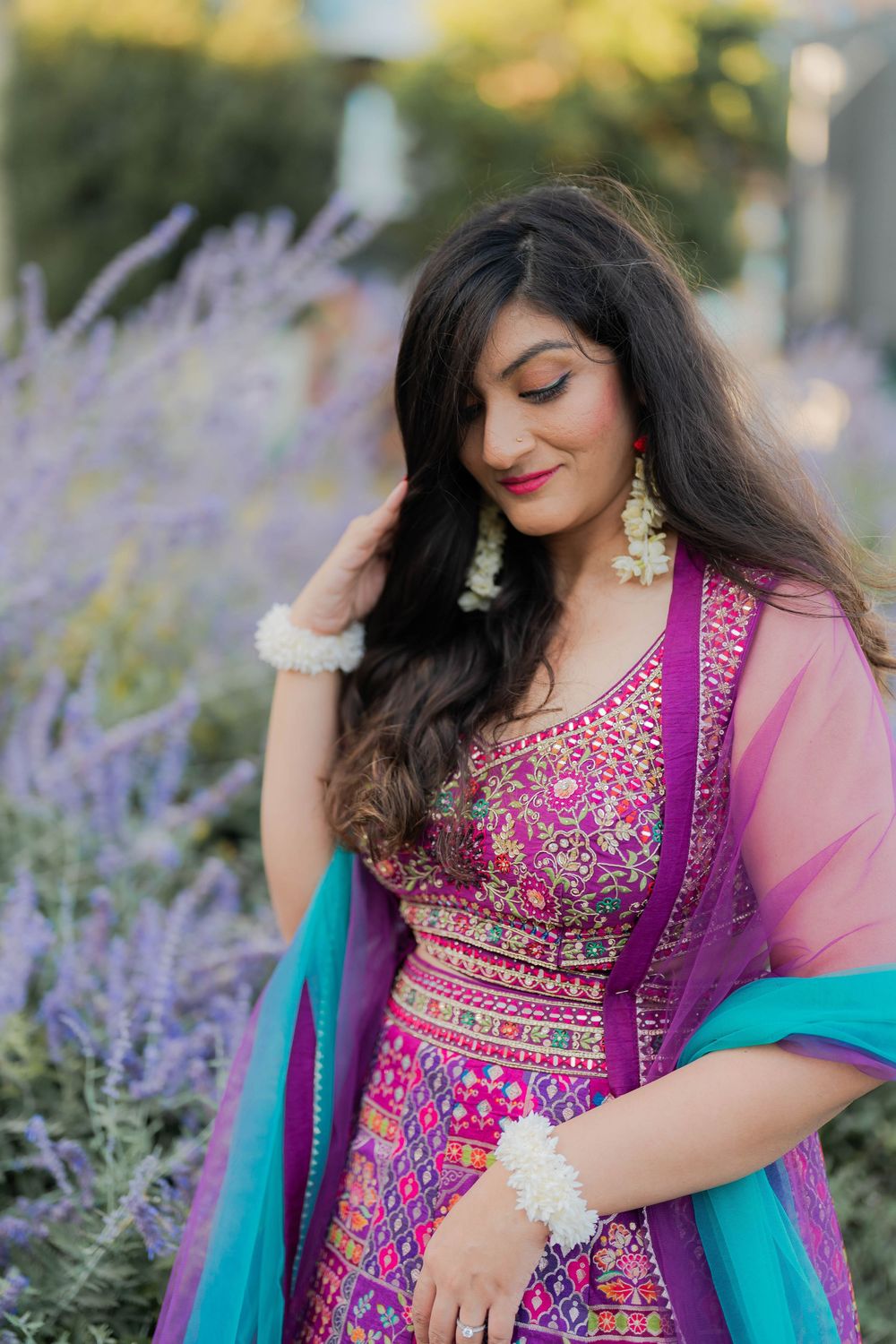 Woman in vibrant pink embroidered lehenga with teal dupatta posing among purple lavender flowers.