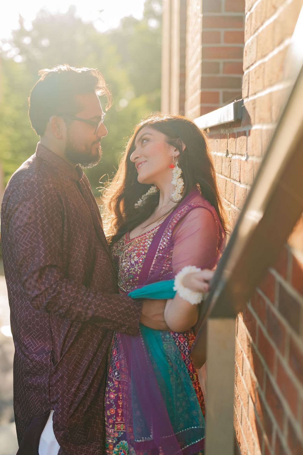 Couple in traditional South Asian attire sharing a romantic moment by a brick wall in golden sunlight.