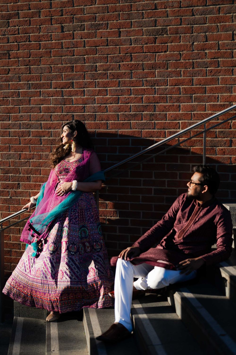 Couple in traditional Indian attire pose on steps against brick wall, woman in pink lehenga, man in maroon kurta.