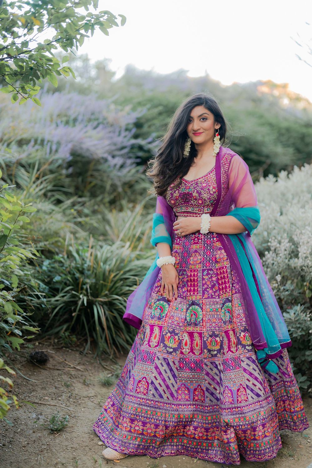 Woman in vibrant purple embroidered lehenga with teal dupatta posing outdoors among lush greenery.