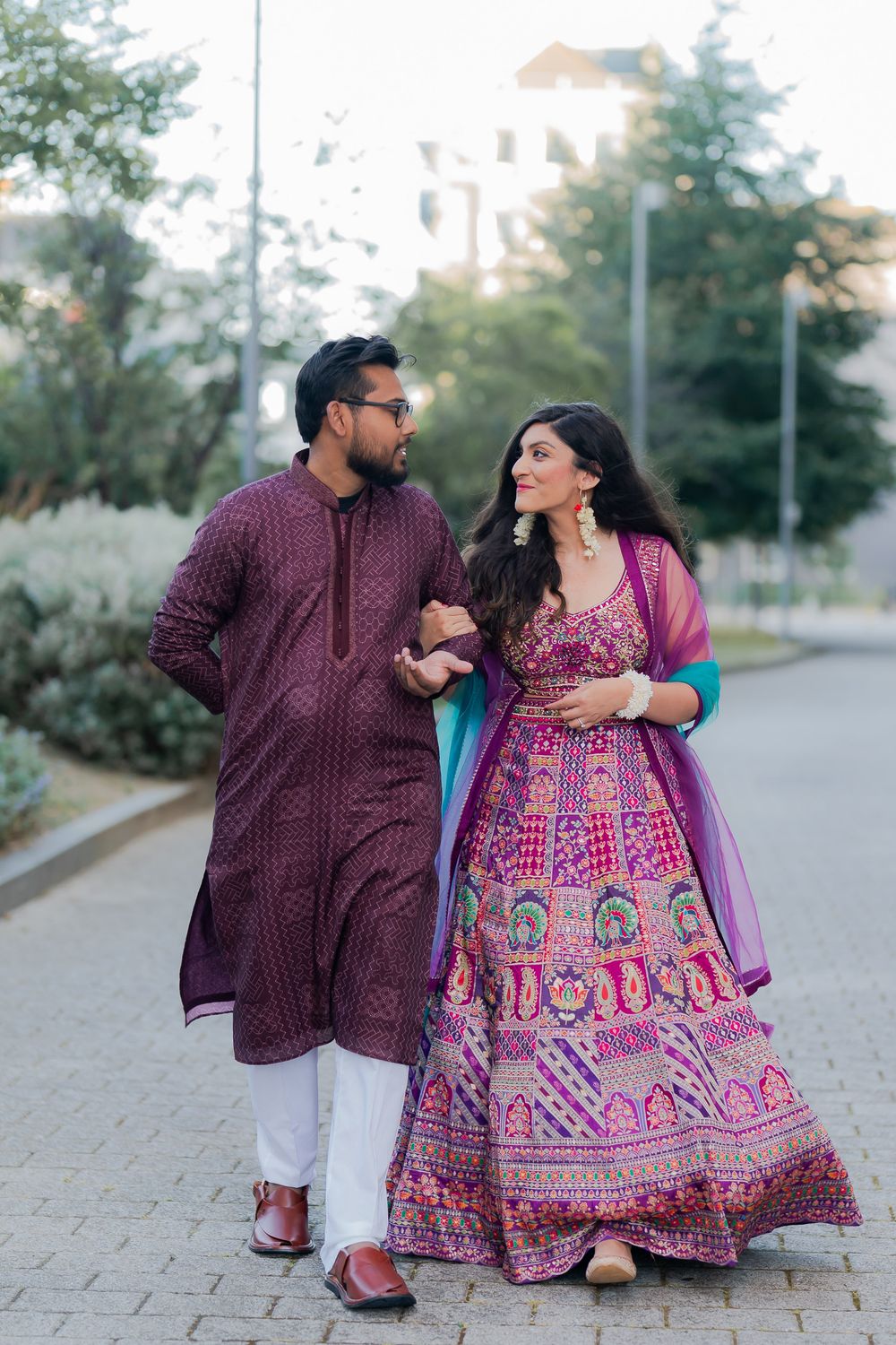 Couple in coordinating purple Indian attire walking outdoors, woman in embroidered lehenga, man in kurta.