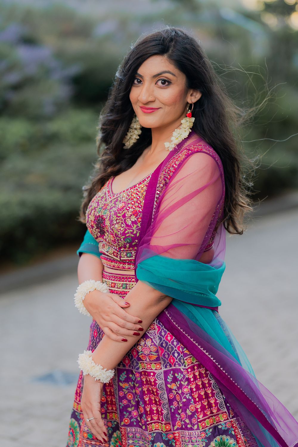 Woman in vibrant pink and teal Indian lehenga with floral jewelry, posing gracefully outdoors.