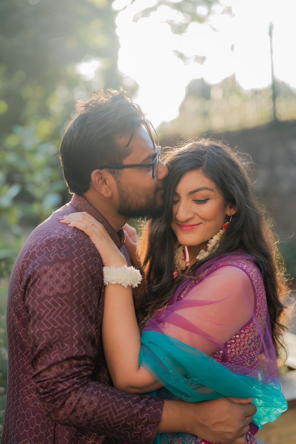 A couple in traditional Indian attire sharing a romantic moment outdoors, man kissing woman on cheek in golden sunlight.