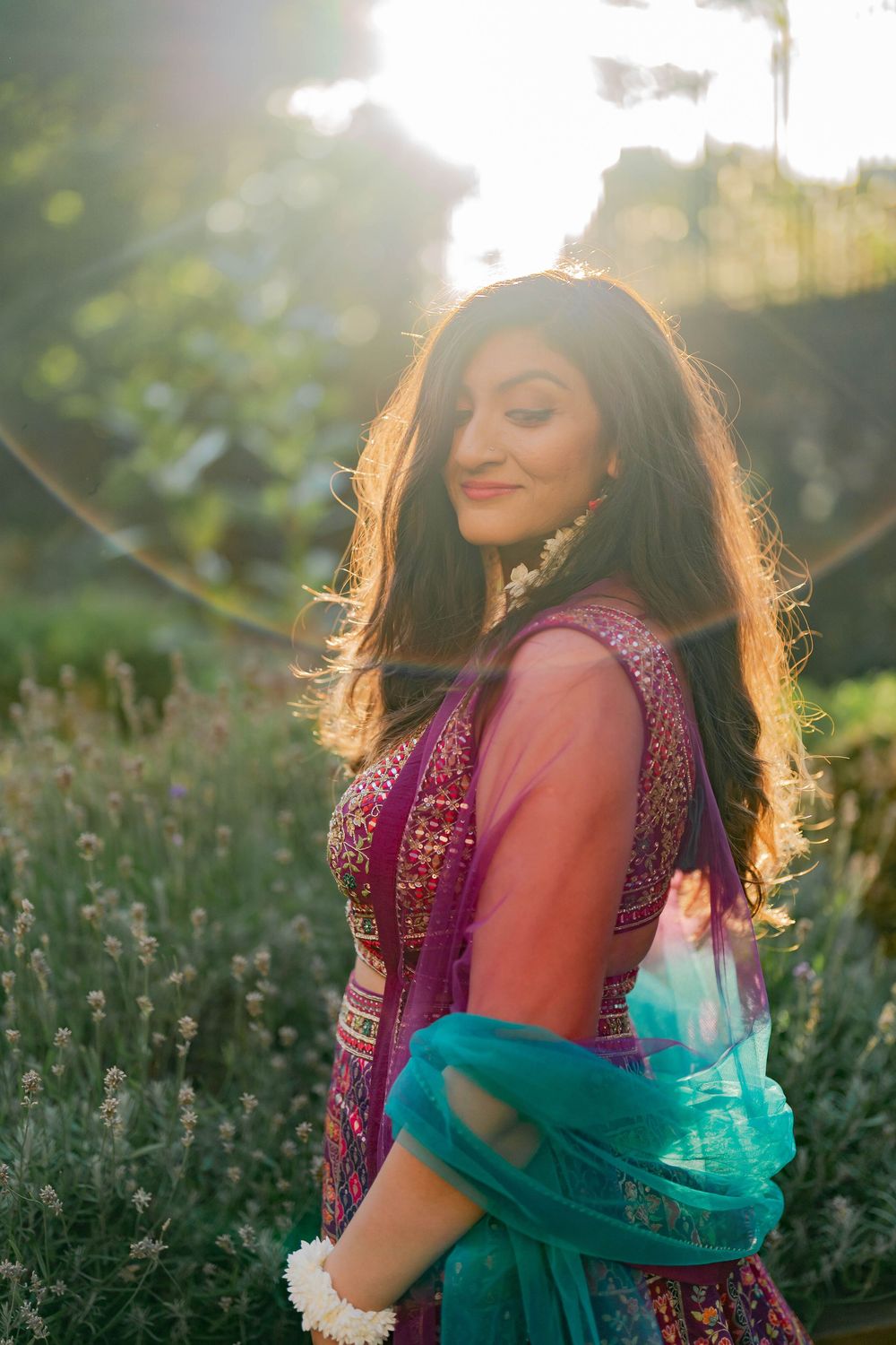 Woman in vibrant pink Indian attire with teal dupatta, smiling outdoors in golden sunlight among lavender plants.