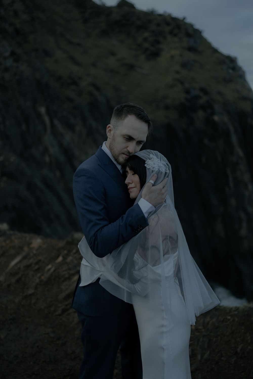 Couple embracing in wedding attire with dramatic rocky cliffs backdrop, bride in veil and groom in navy suit.