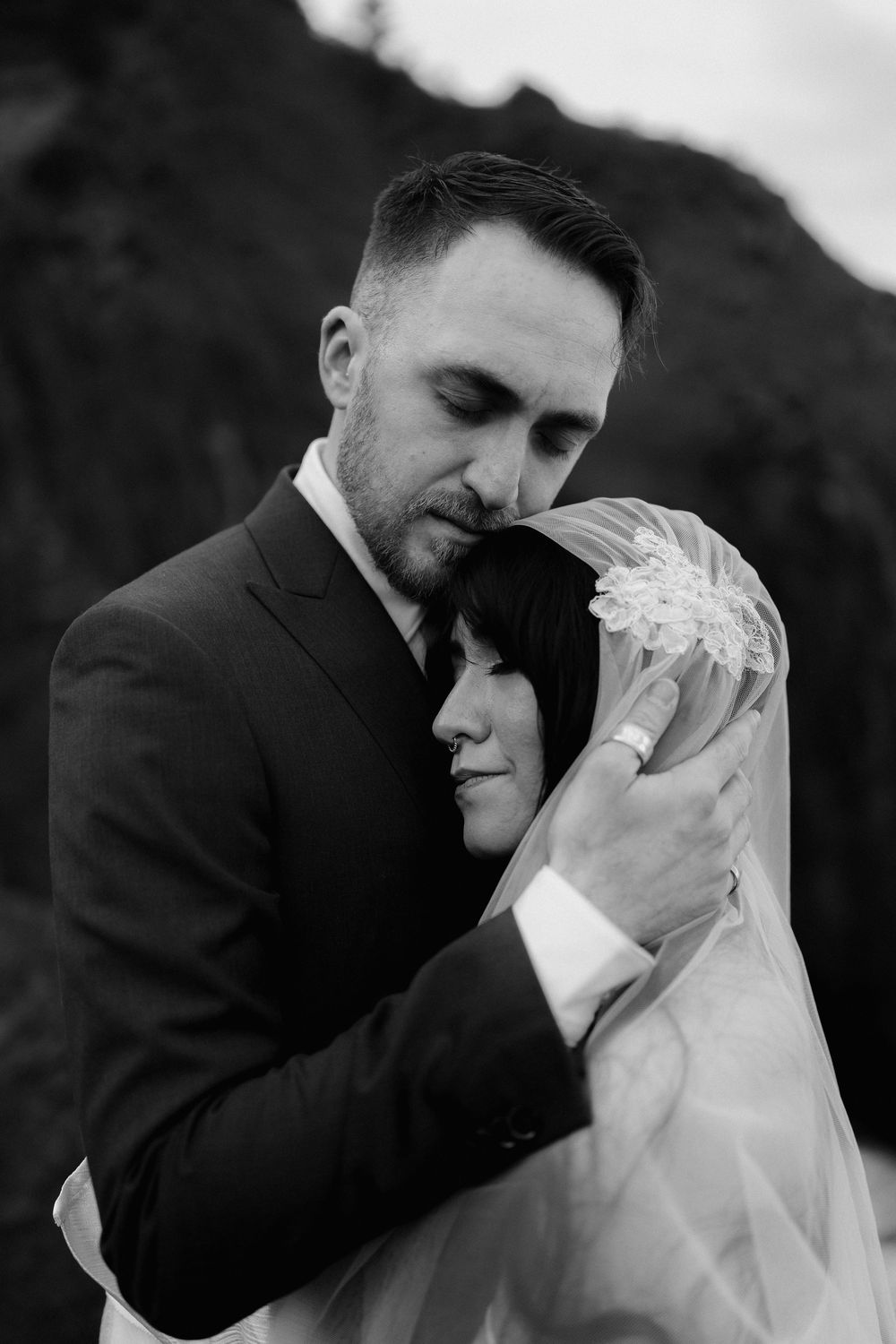 Black and white wedding portrait of groom embracing bride in veil, sharing an intimate moment outdoors.