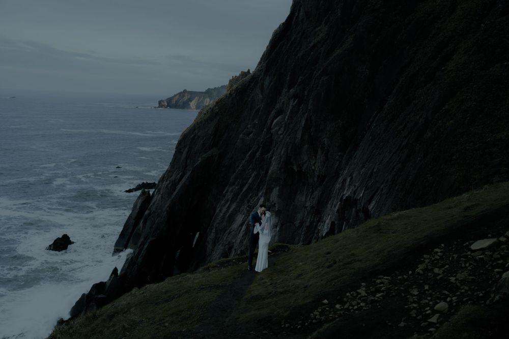 Couple in white wedding attire standing on dramatic coastal cliffs overlooking a dark stormy ocean.