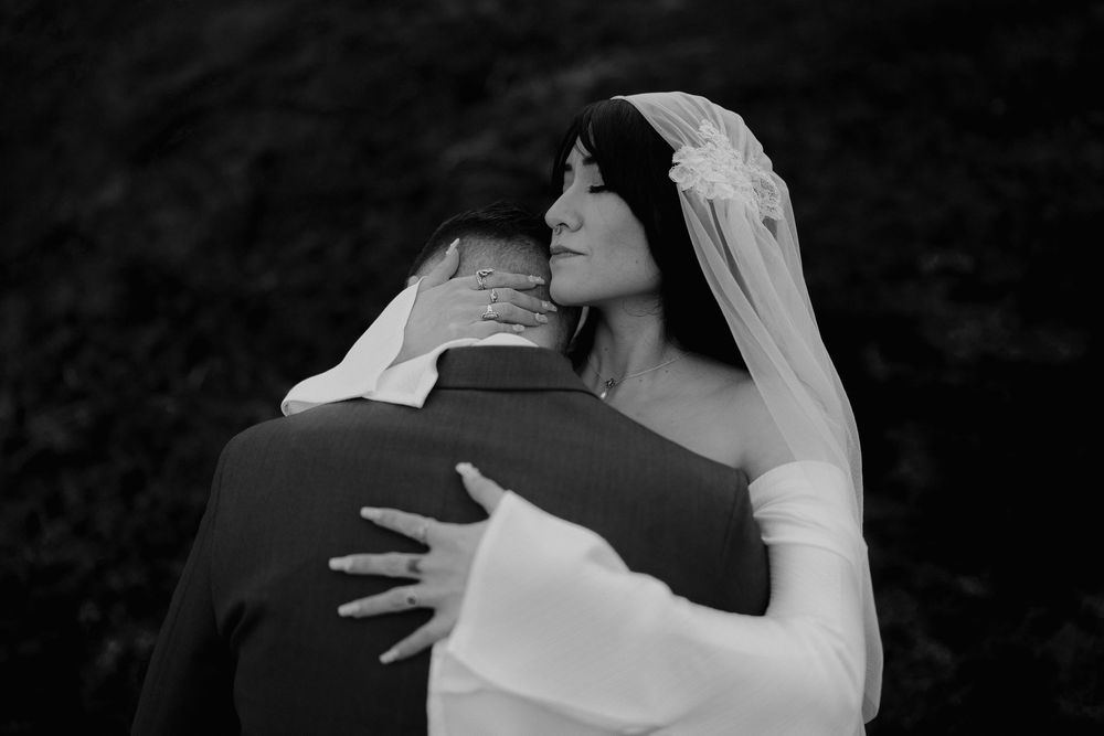 Black and white photo of a bride in a veil embracing a groom from behind in an elegant wedding portrait.