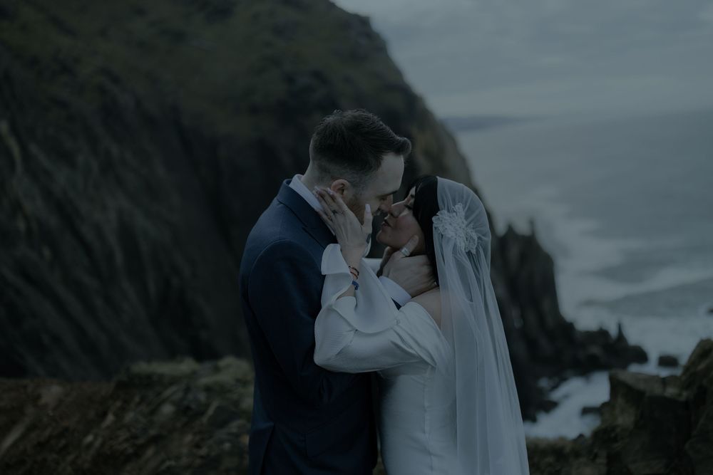 A couple shares an intimate moment at their coastal wedding, with dramatic cliffs and ocean waves in the moody background.