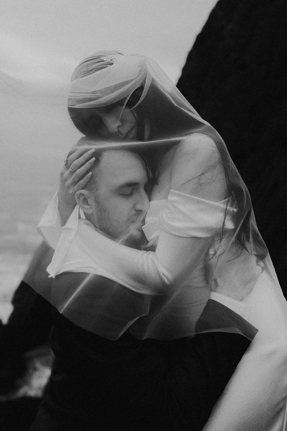 Black and white photo of a bride and groom sharing an intimate embrace under a flowing veil outdoors.