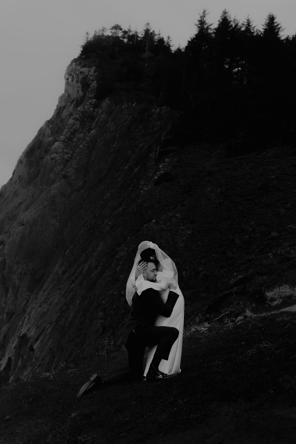 Black and white photo of a couple embracing in wedding attire on a rocky cliffside with pine trees above.