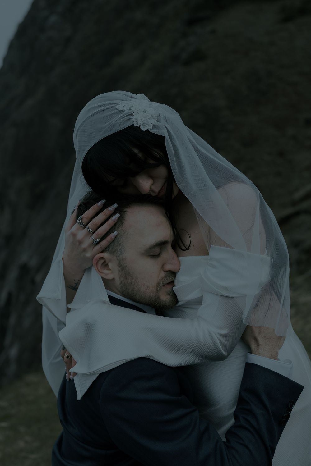 A bride draped in a white veil embraces her groom tenderly outdoors against a dark rocky backdrop.