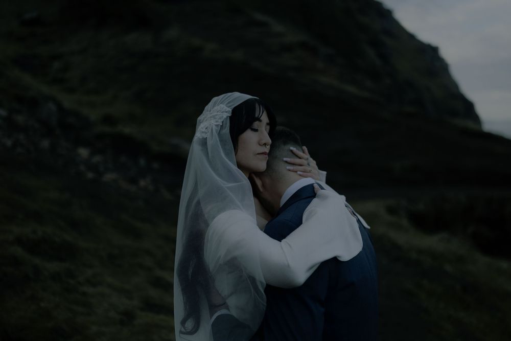 A bride in a white veil embraces a groom on a misty hillside in a romantic outdoor wedding portrait.