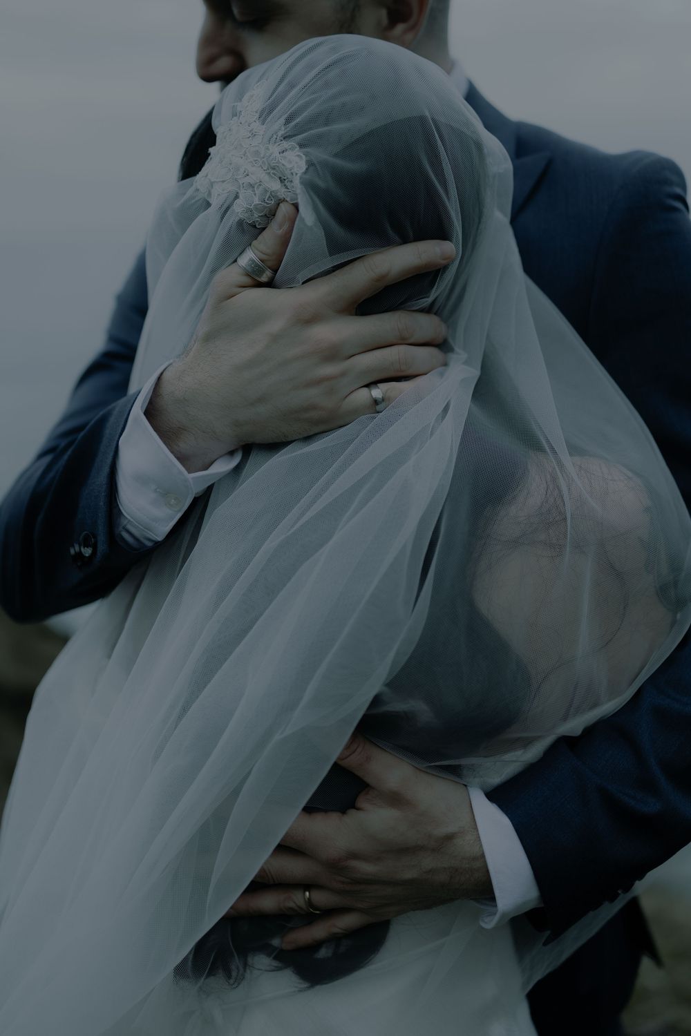 A bride and groom embrace tenderly, her white veil draped over them both, exchanging rings on their wedding day.