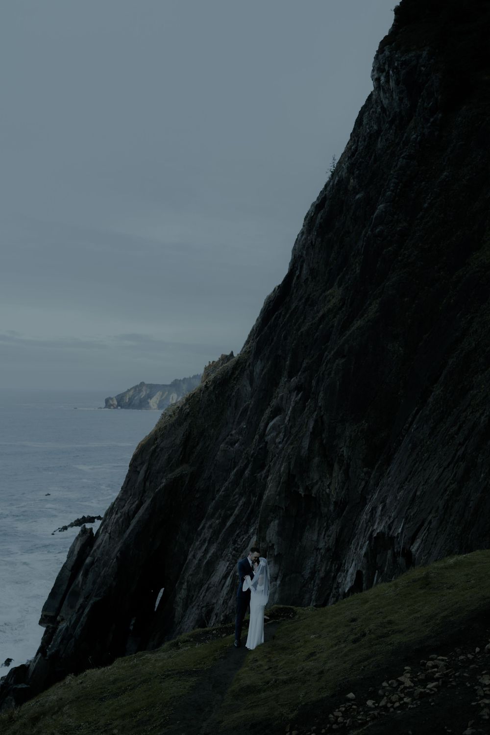 Couple in white embracing on dramatic coastal cliffs overlooking a moody ocean under overcast skies.