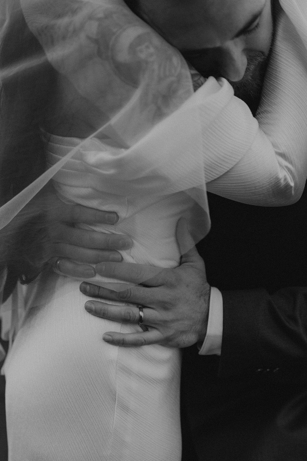 Black and white wedding photo of groom embracing bride, his ringed hand resting on her waist beneath a flowing veil.