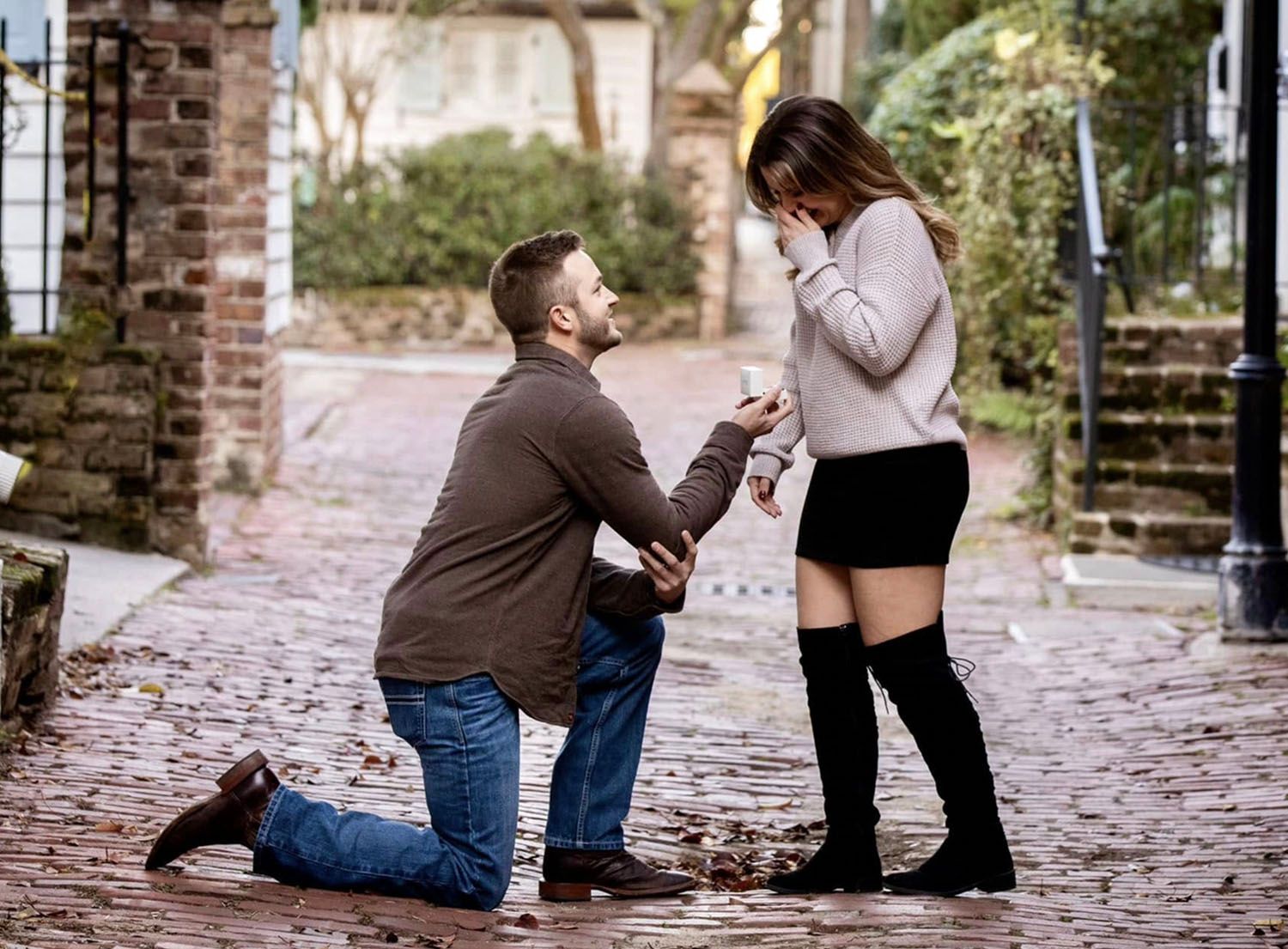 Man proposing to woman on a cobblestone street, holding a ring box as she reacts with surprise and joy.