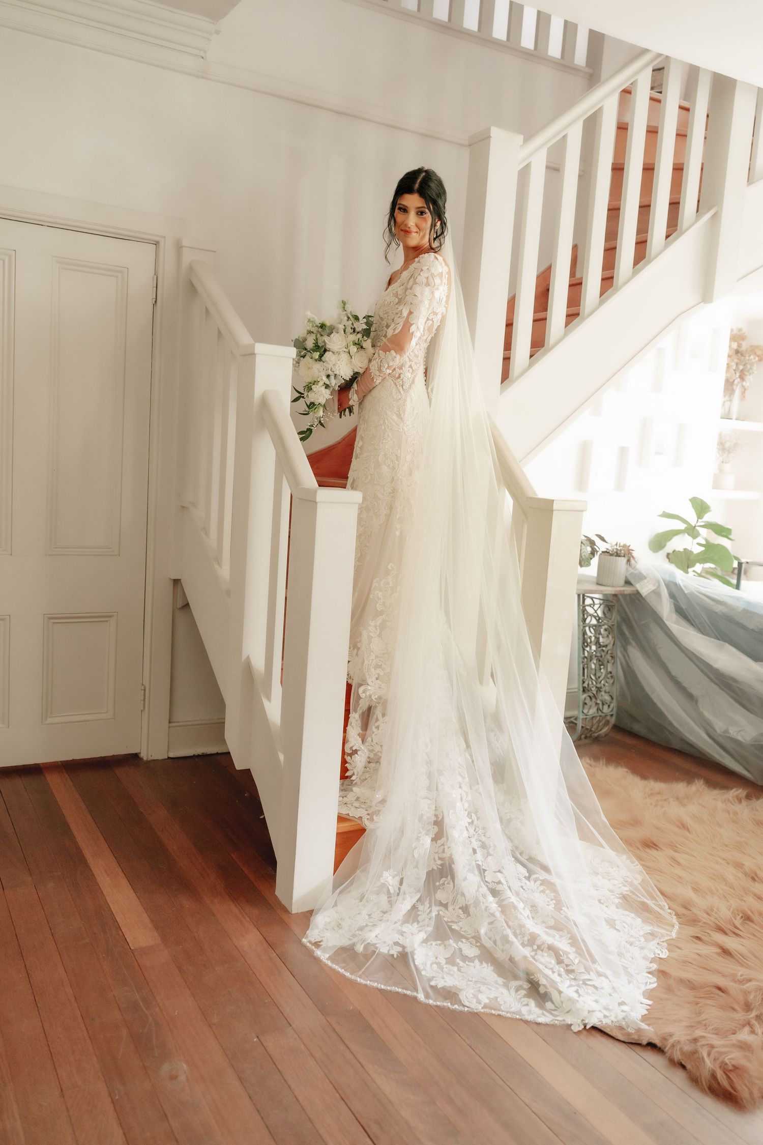 Bride in lace wedding gown with cathedral veil stands on staircase in elegant white interior.