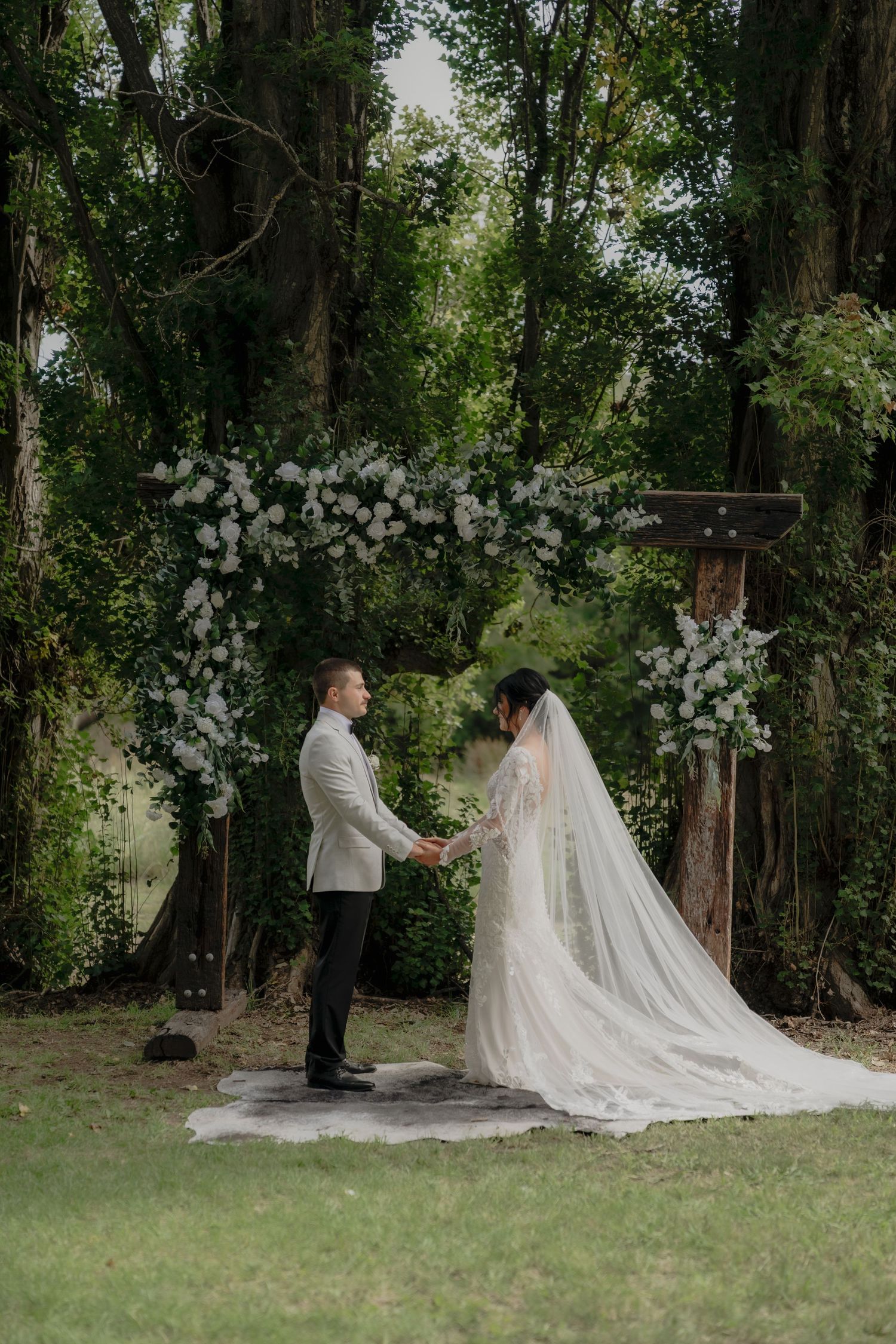 Couple exchanges vows outdoors under floral arch surrounded by lush green trees.