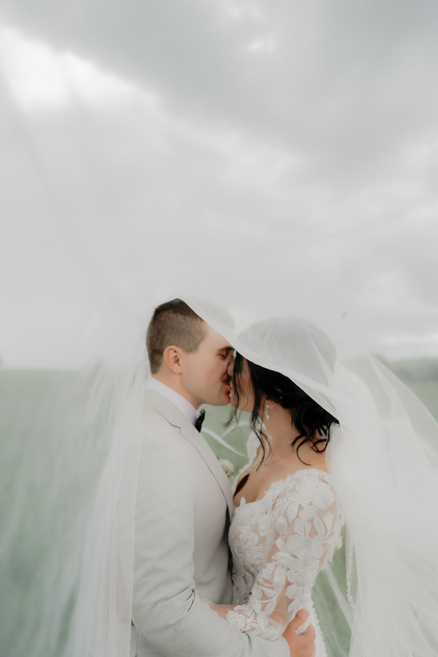 Bride and groom share a romantic kiss under a flowing white veil on their wedding day with cloudy skies.