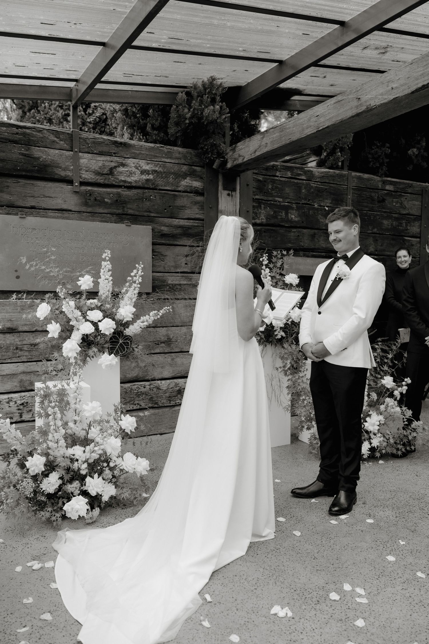 Black and white photo of bride and groom exchanging vows at outdoor wedding ceremony with floral arrangements.