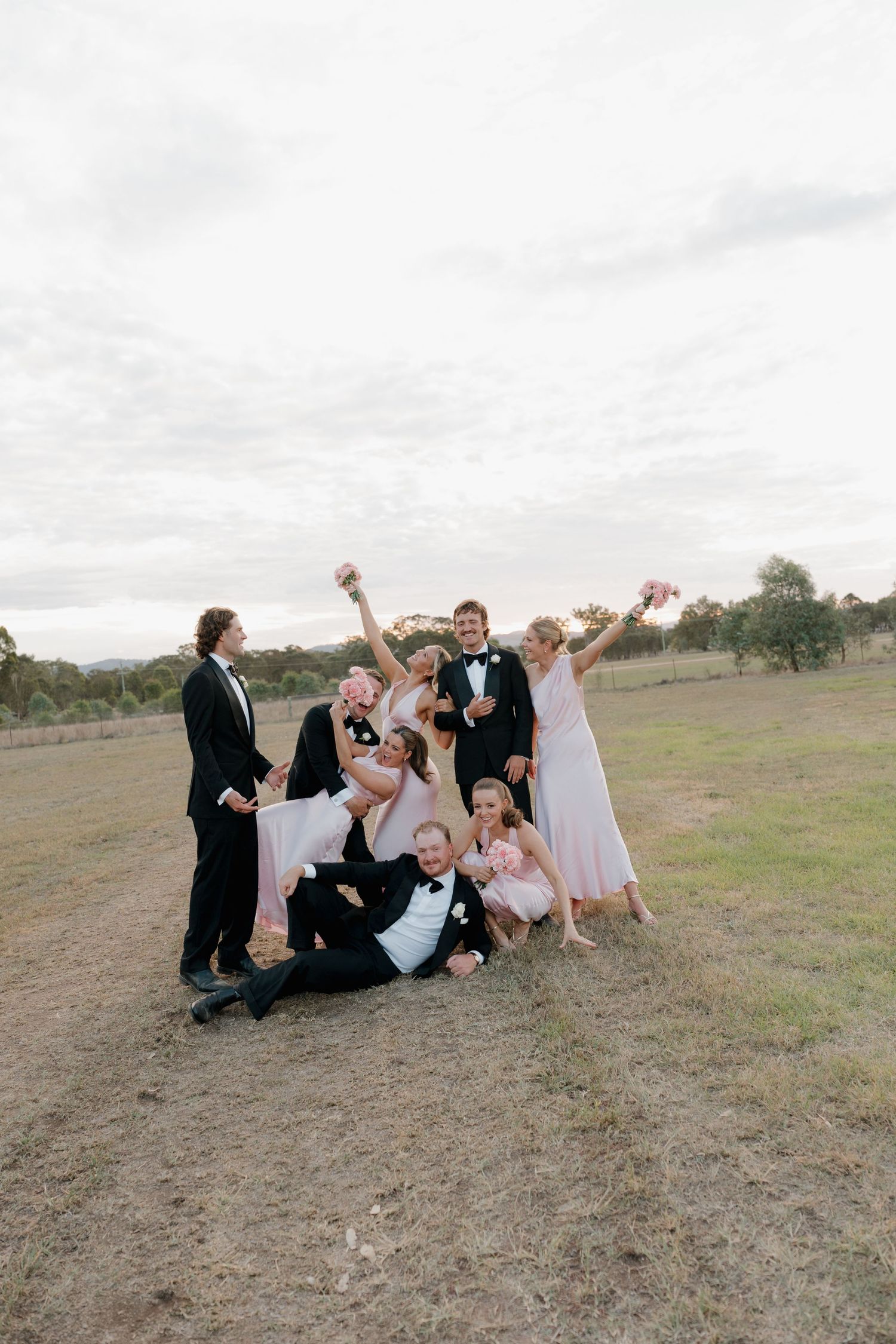 Wedding party celebrating outdoors in formal attire, bridesmaids in pink dresses and groomsmen in tuxedos, posing joyfully in a field.