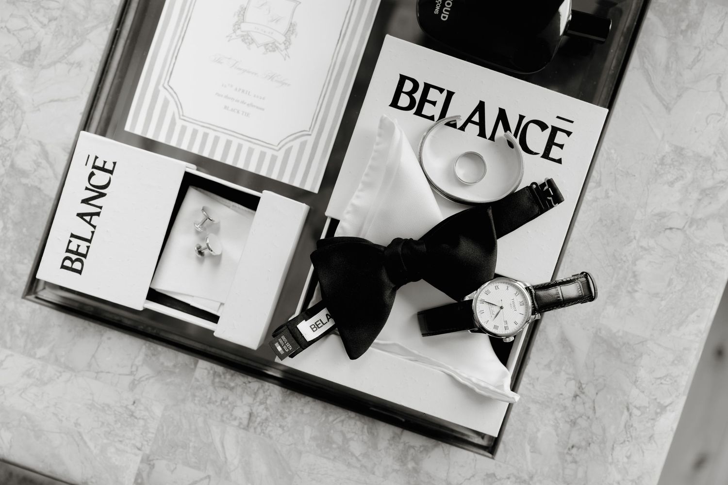 Black and white flat lay of Belance watch, bow tie, cufflinks, and wedding invitation on marble surface.