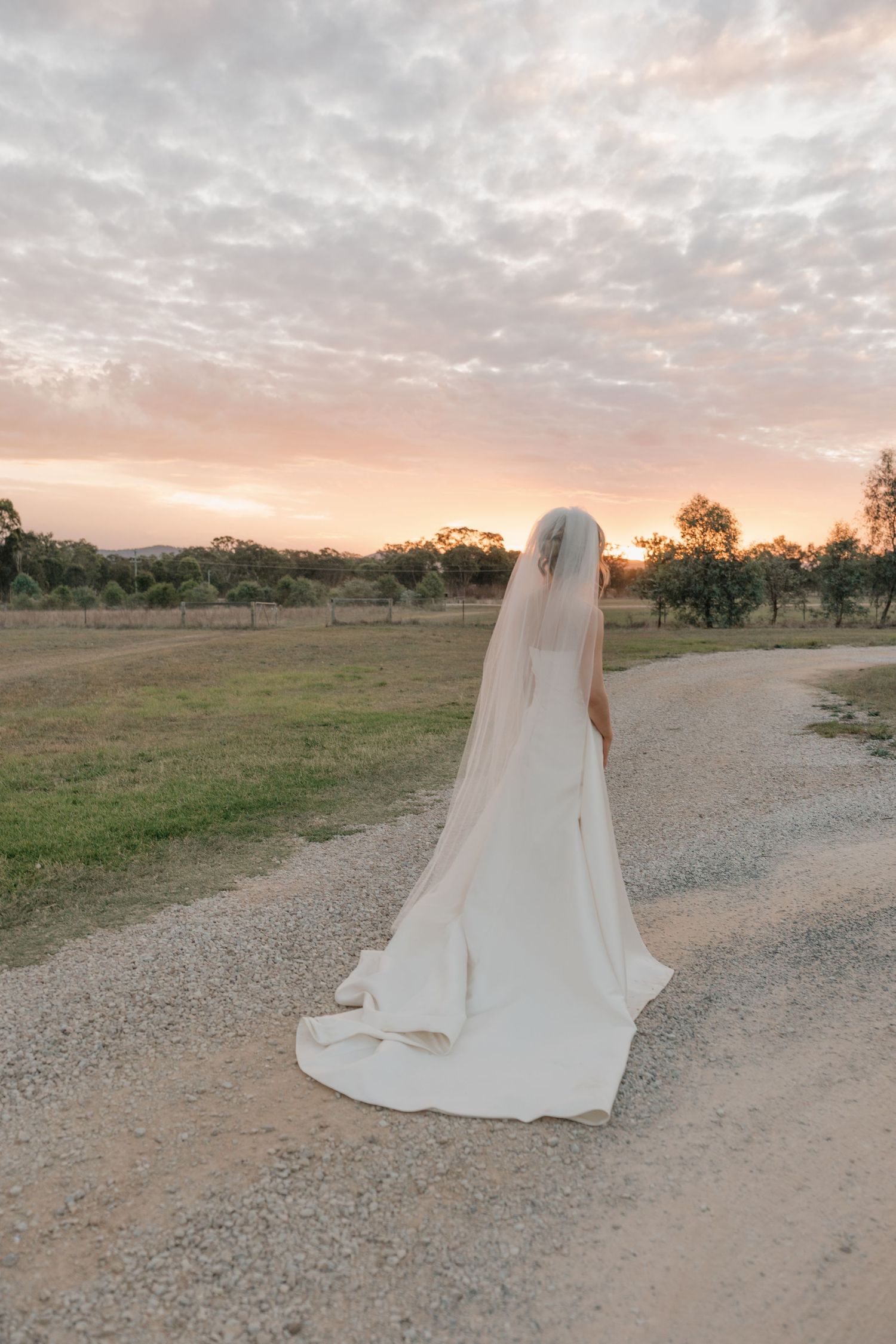 Bride in white gown and veil stands on gravel path at sunset, overlooking scenic countryside landscape.