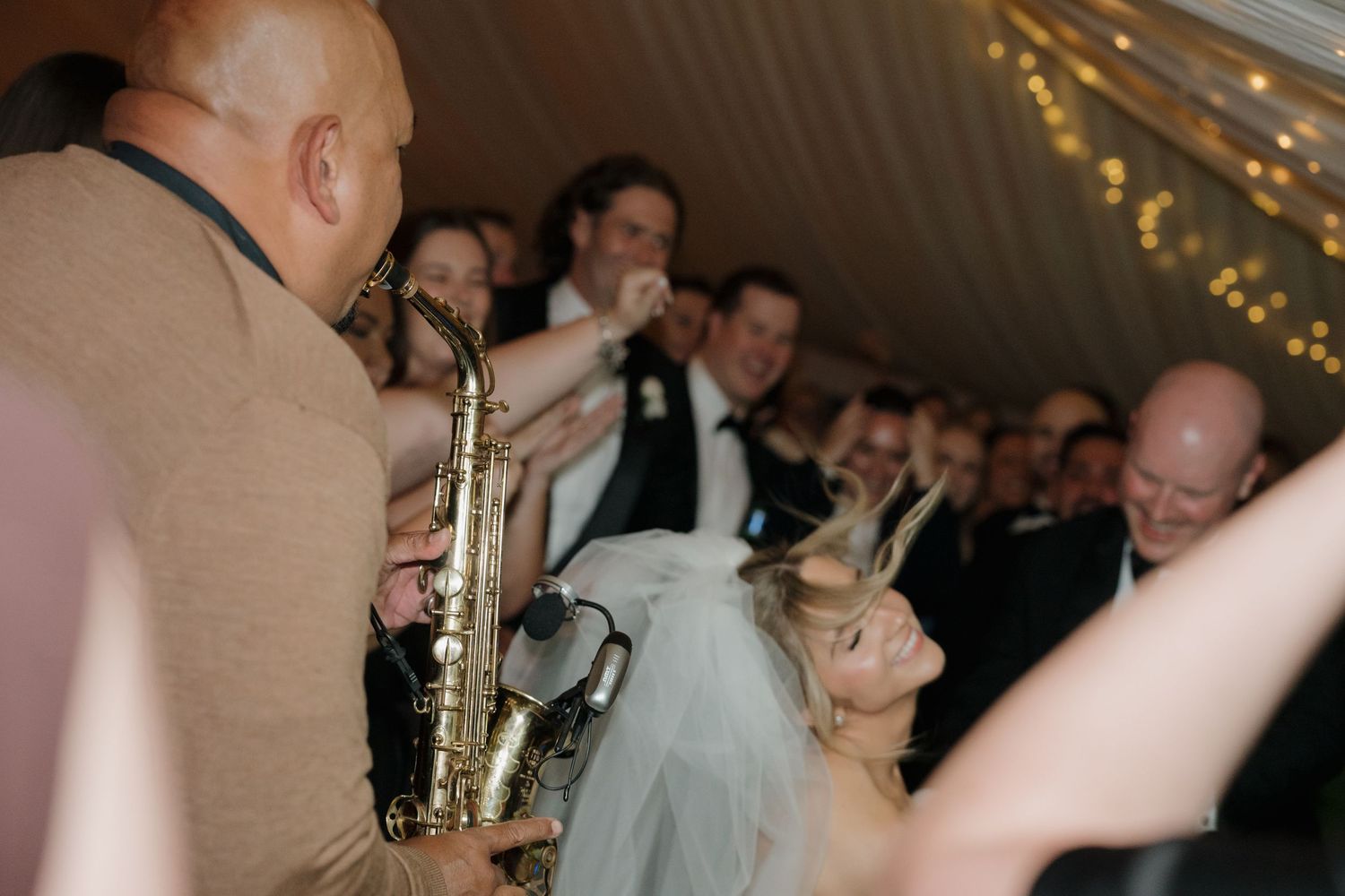 Saxophonist performing for bride and groom surrounded by wedding guests at elegant tented reception.