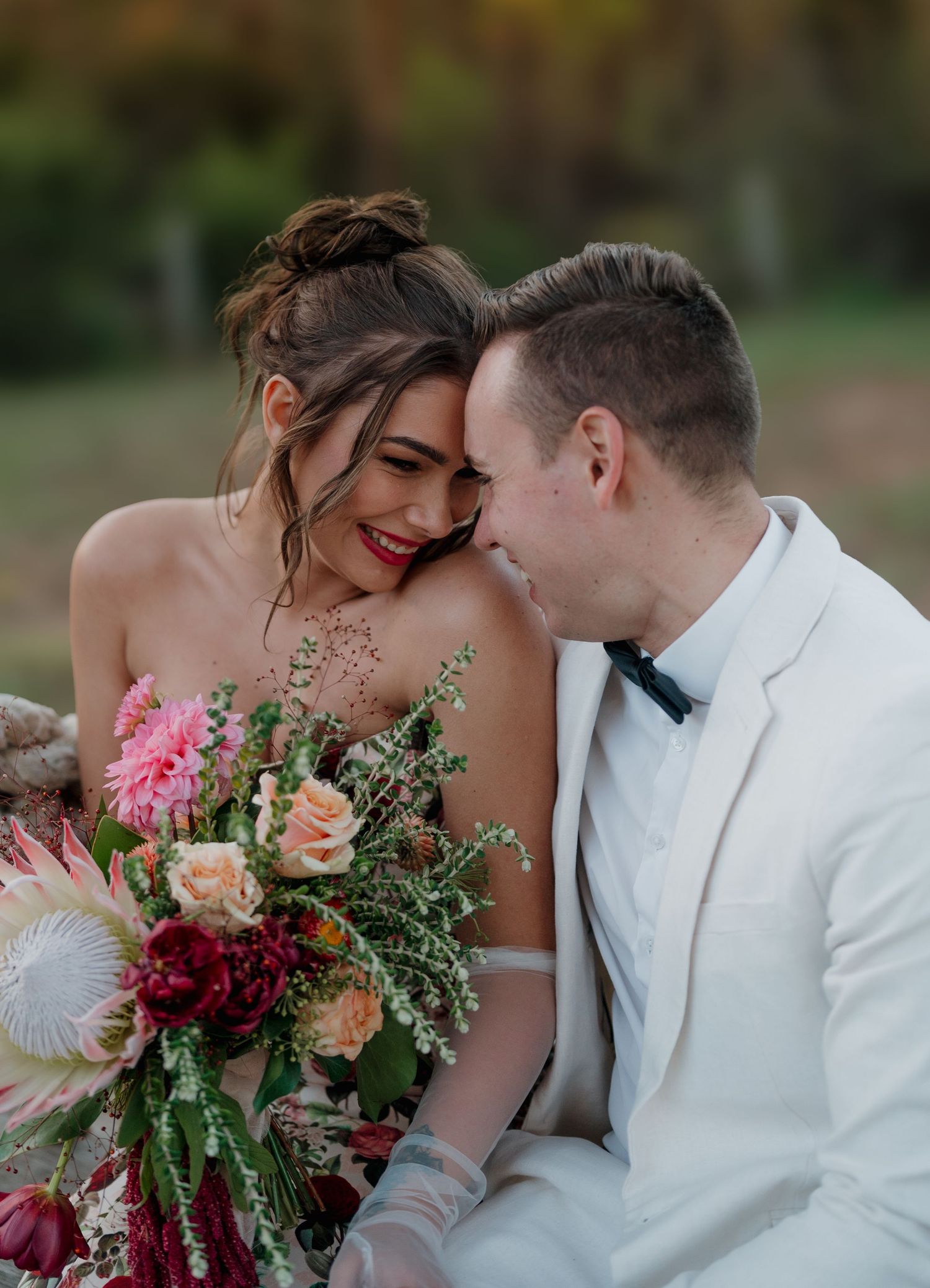 Bride and groom share an intimate moment, she holds a vibrant pink and red floral bouquet outdoors.