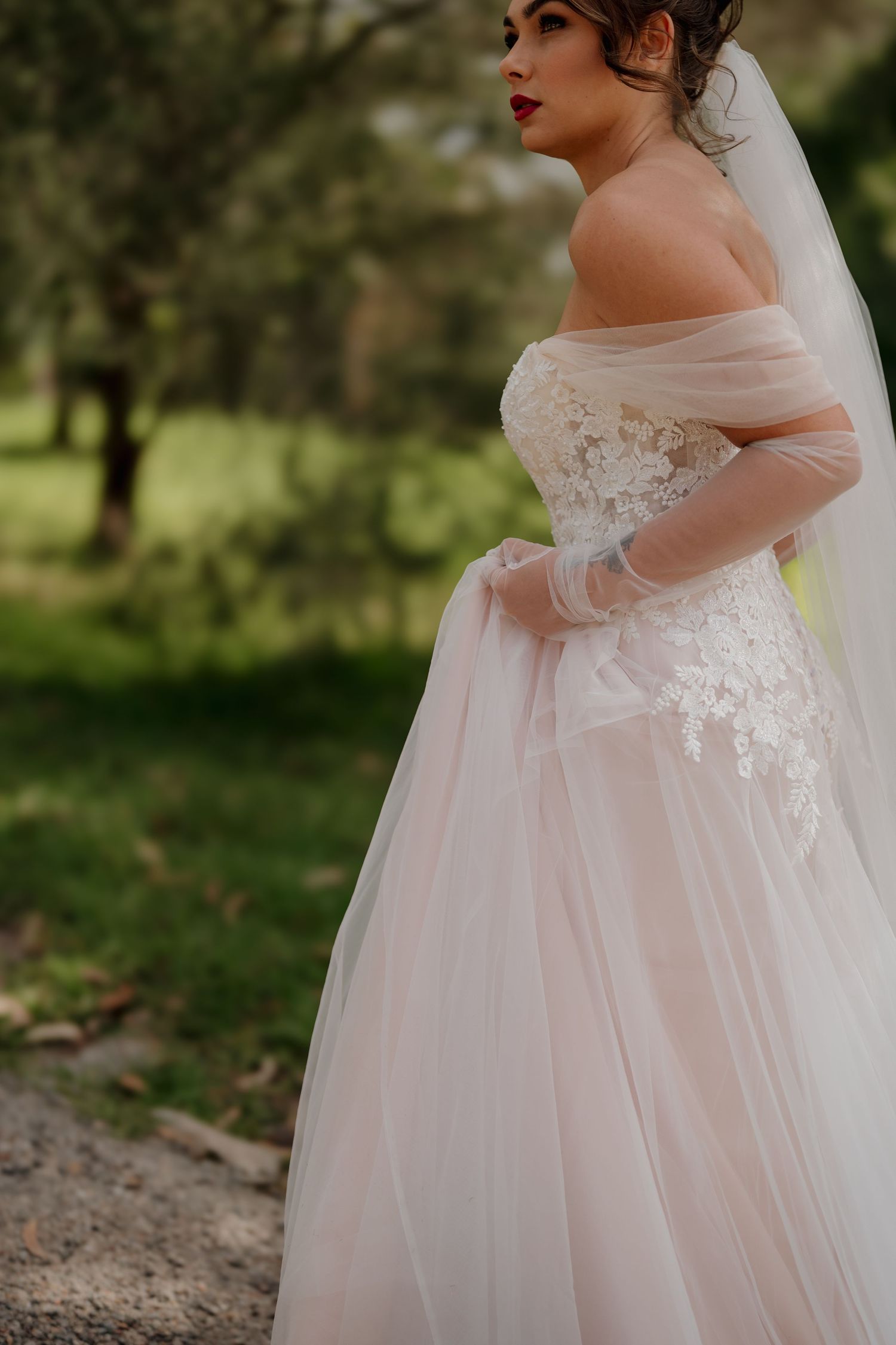 Bride in off-shoulder blush tulle wedding gown with lace bodice and veil, posing outdoors among green trees.