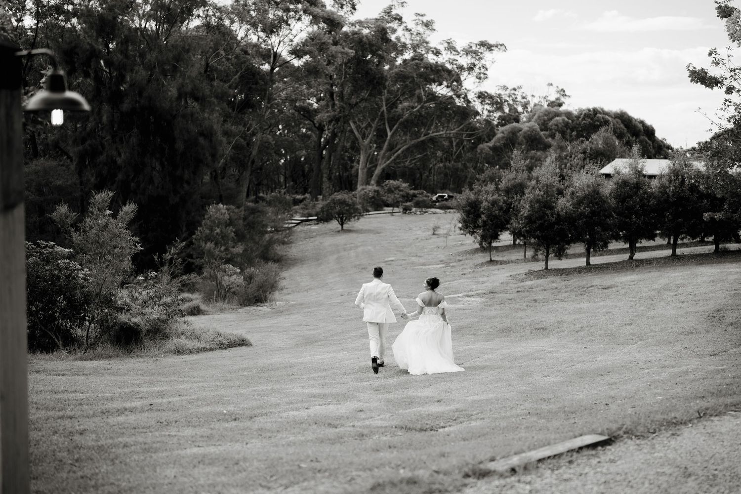 Black and white photo of a bride and groom walking hand in hand across a lush green lawn surrounded by trees.