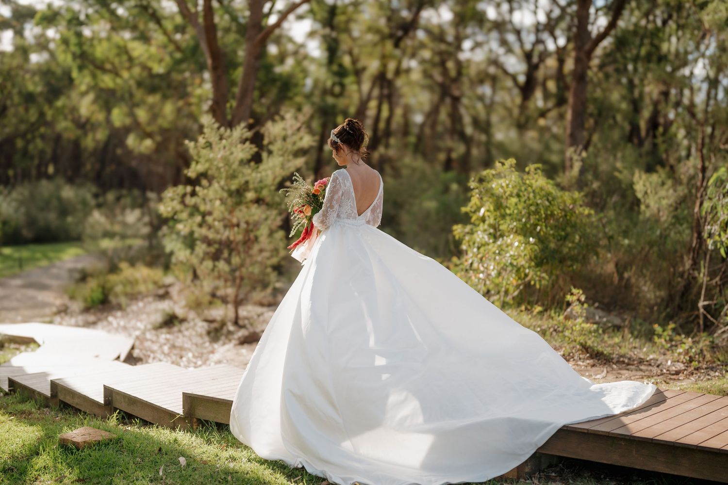 Bride in a flowing white wedding gown with a long train stands on a wooden path surrounded by lush green trees.