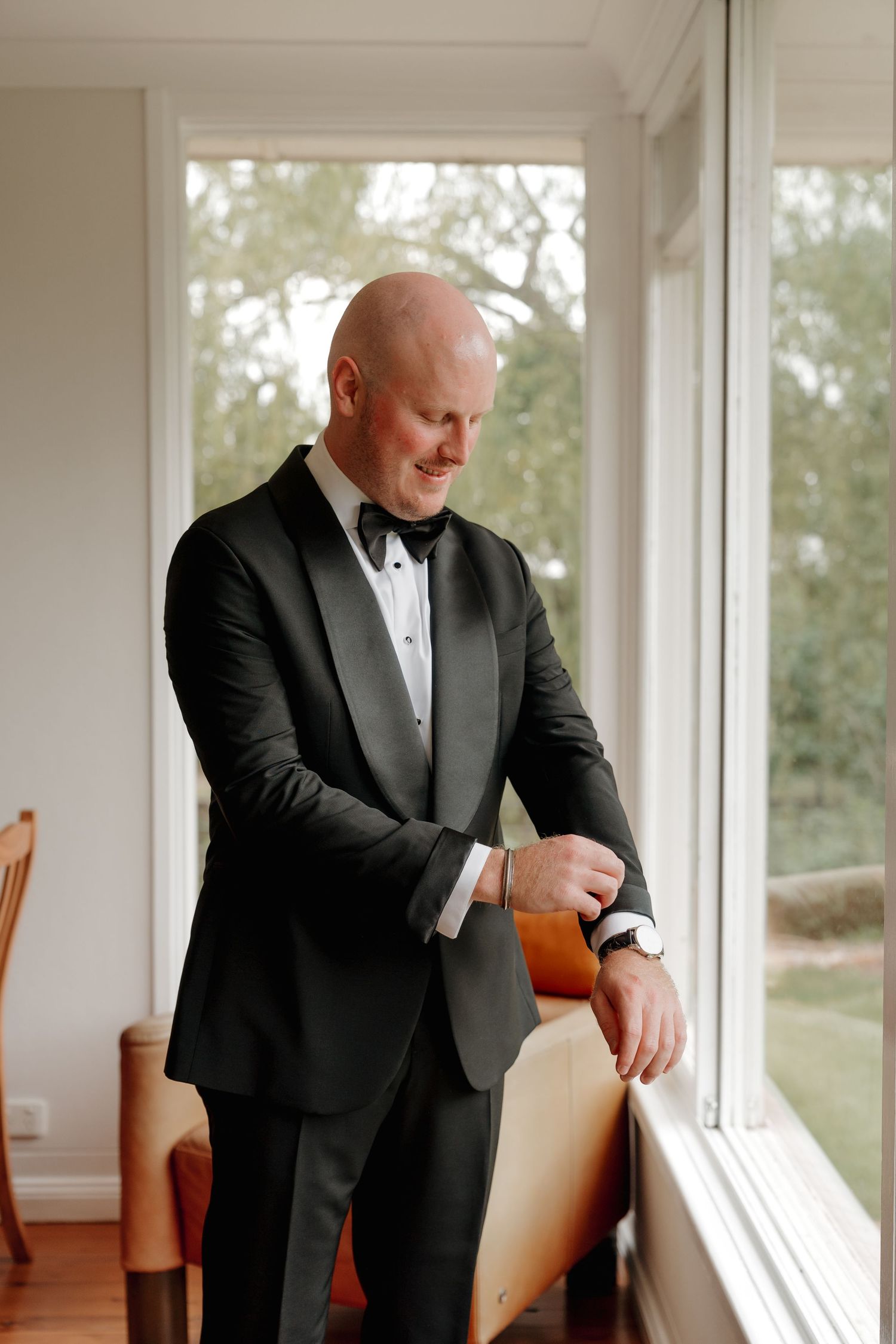 Bald groom in black tuxedo and bow tie adjusting cufflinks by a sunlit window.