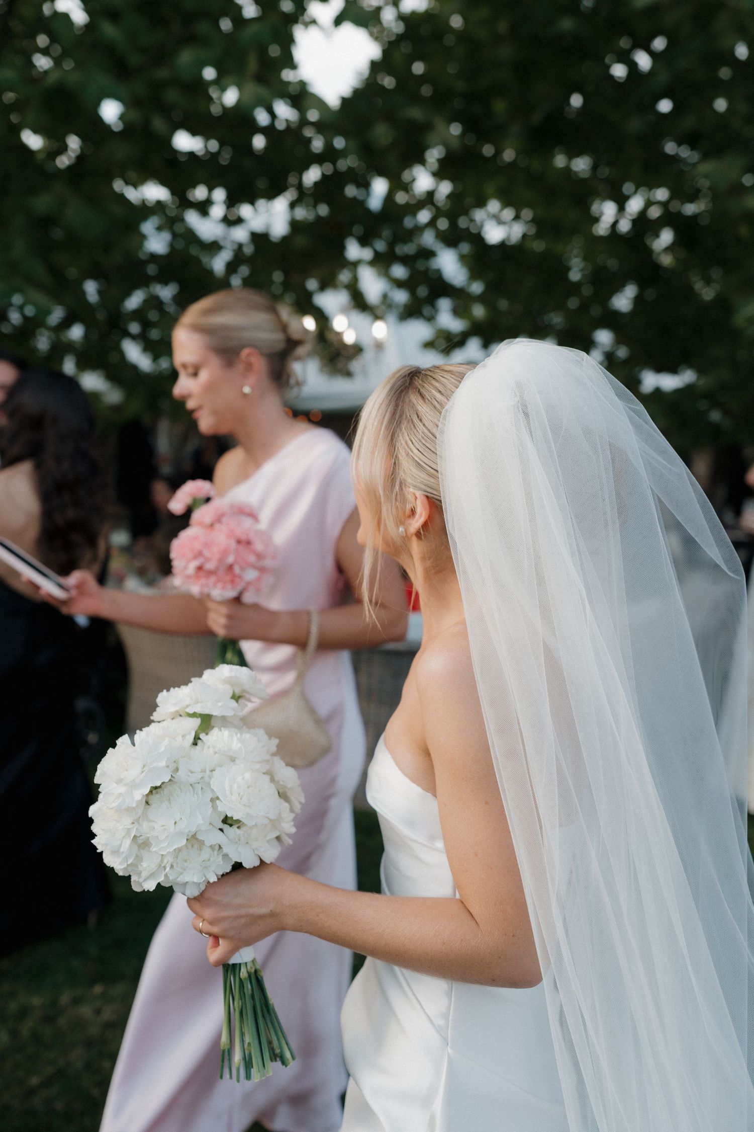 Bride in white gown and veil holds white bouquet, with bridesmaid in pink dress carrying pink flowers outdoors.