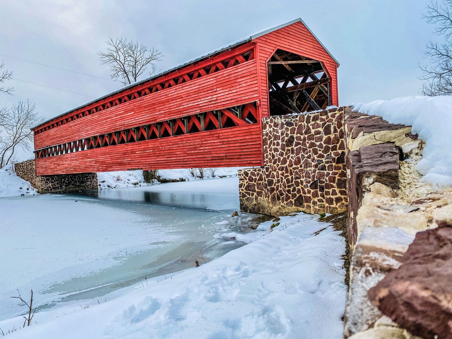 Red covered bridge over frozen river in winter snow landscape with stone foundation.