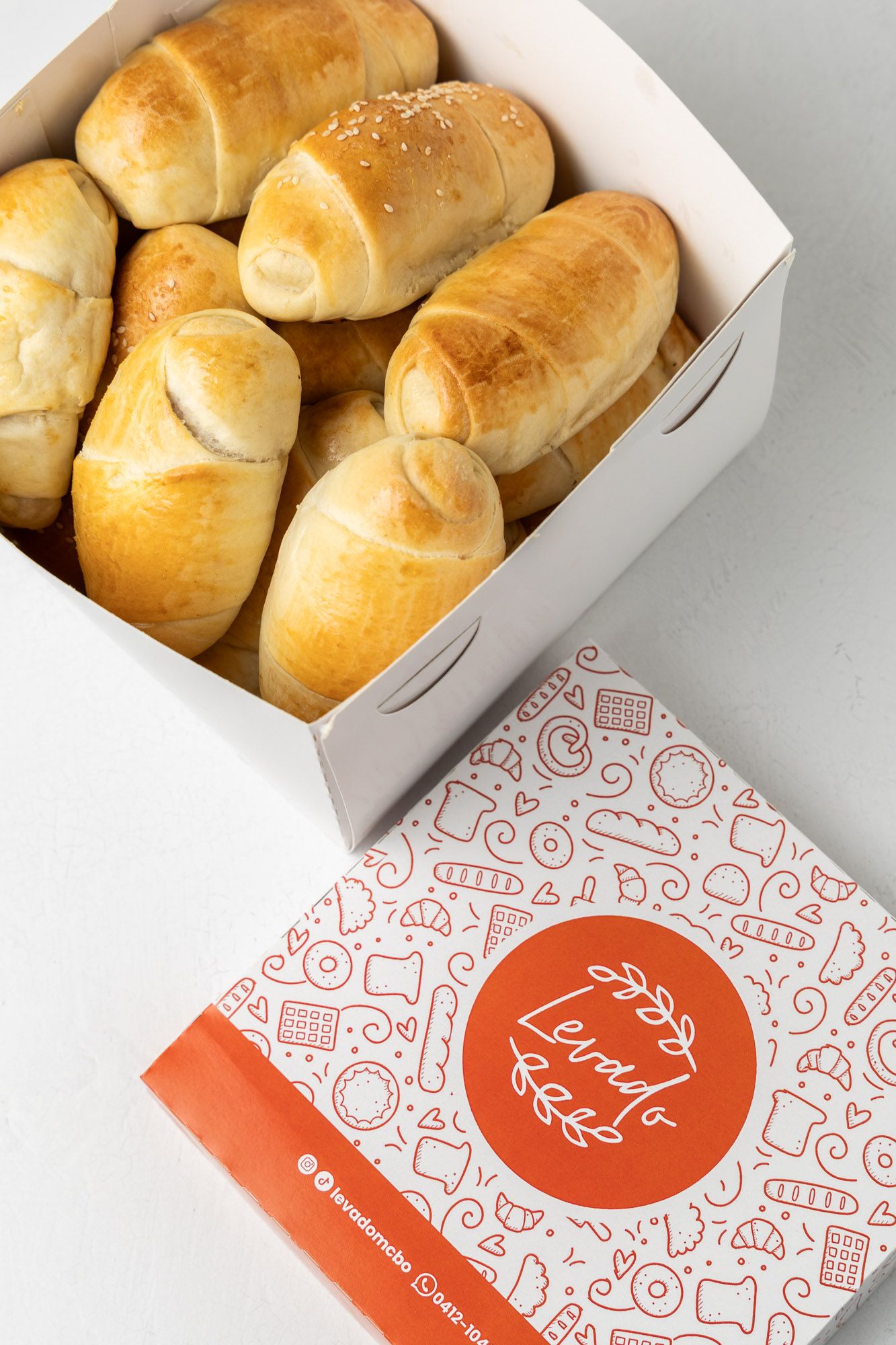 Freshly baked golden bread rolls in a white bakery box next to a decorative Levdig Bakery branded packaging.
