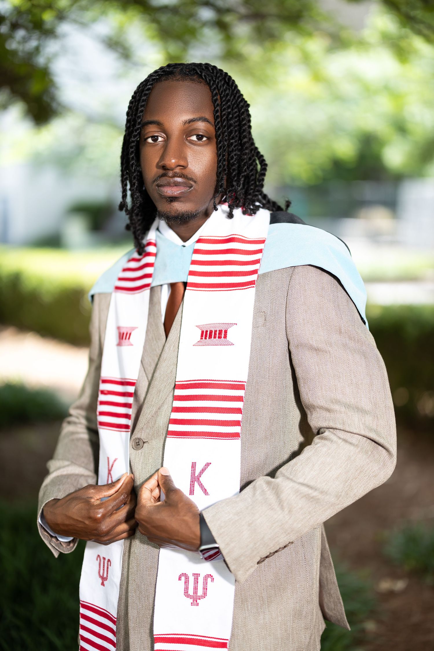 Young Black male graduate in beige suit wearing Indiana University stole with red stripes outdoors.