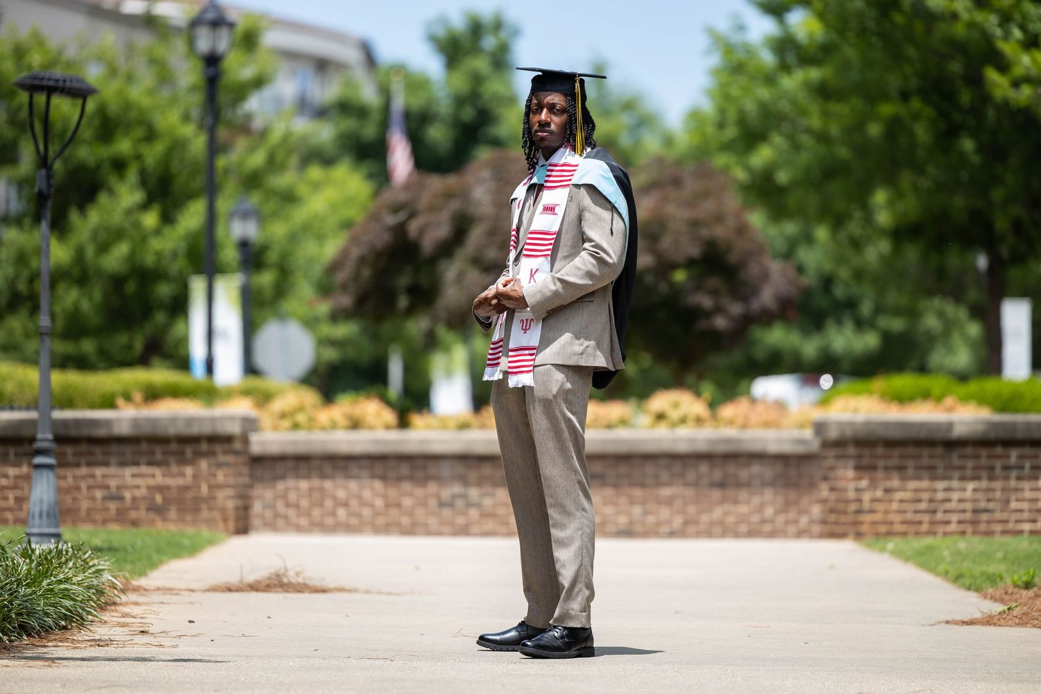 Graduate in cap and gown with stole stands proudly on campus pathway surrounded by lush green trees on a sunny day.