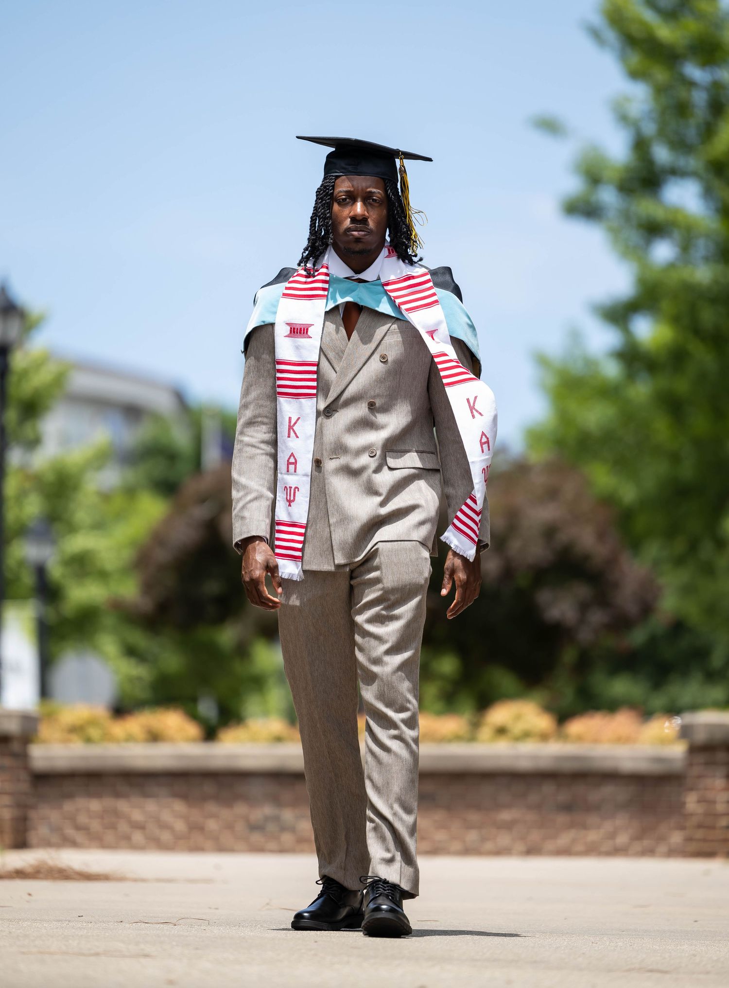Young man in graduation cap, kente stole, and tan suit walks confidently outdoors on a sunny day.