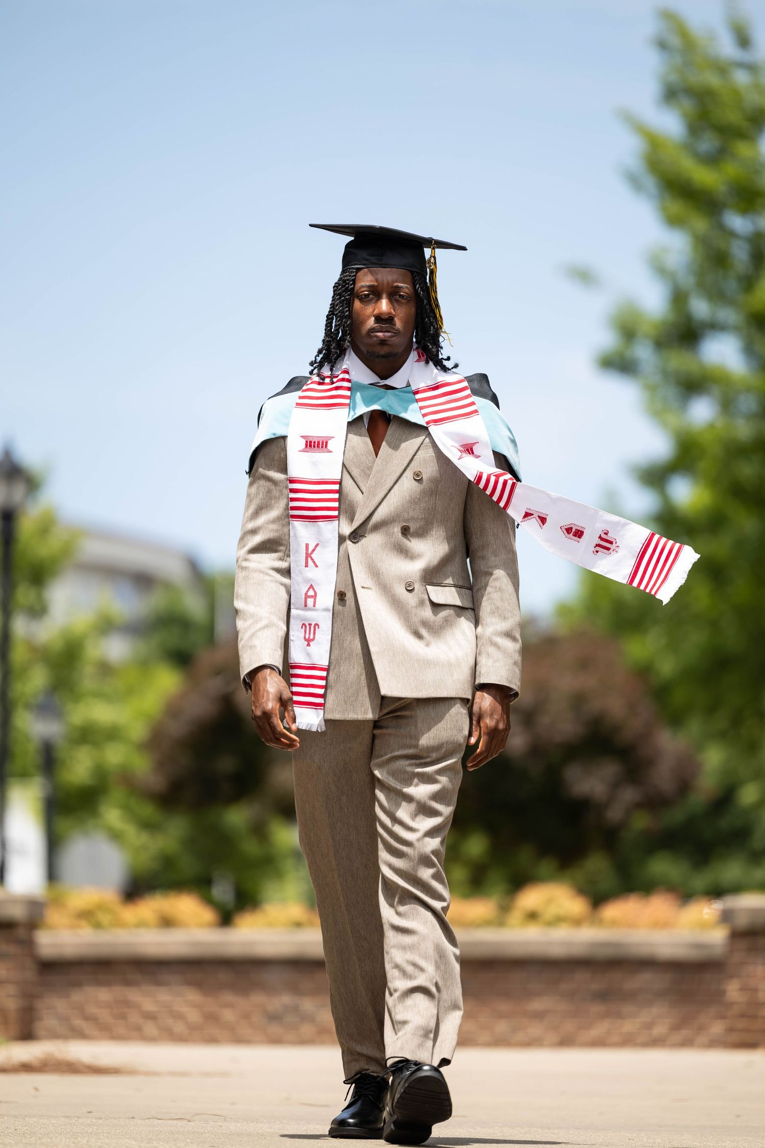 Confident graduate walking in tan suit, cap, and Kappa Alpha Psi stole on sunny campus.