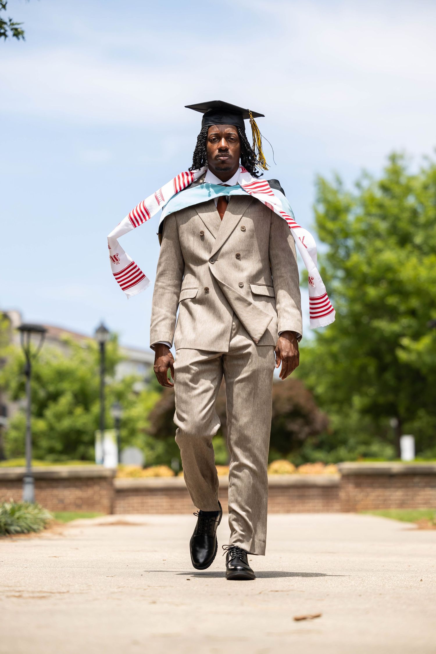 Young Black man in beige suit and graduation cap walks confidently outdoors wearing colorful kente stole on sunny day.
