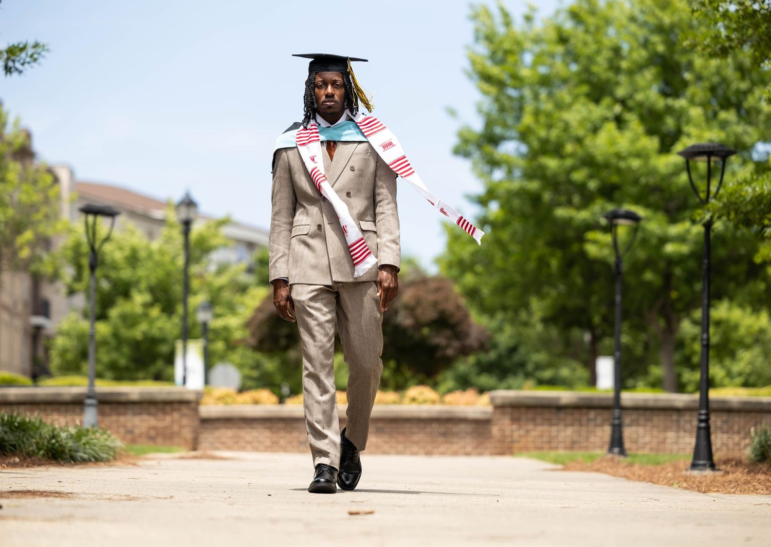 Graduate in cap and gown with American flags walks confidently on a sunny campus path in a stylish suit.