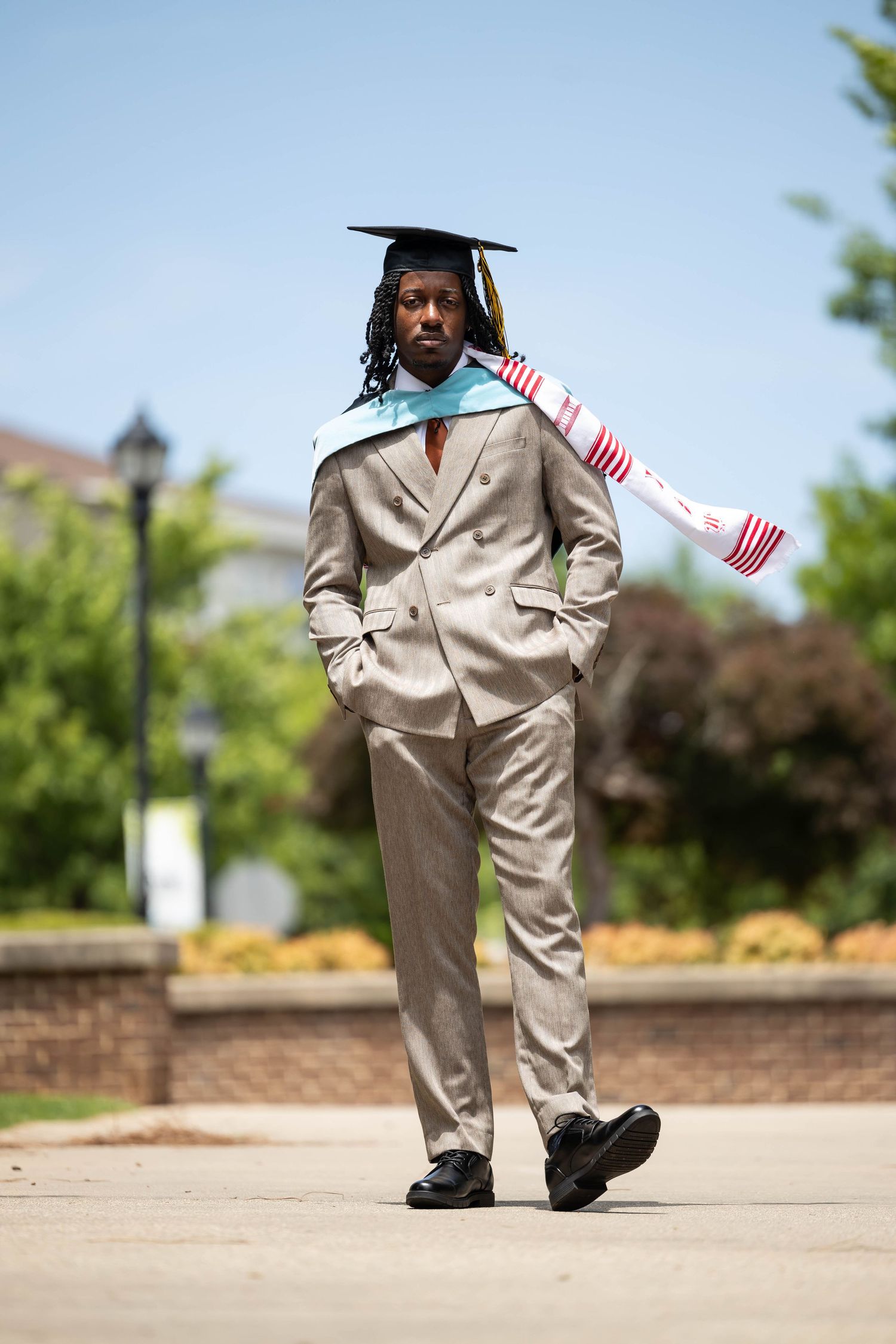 Young Black male graduate in gray suit and mortarboard cap walks outdoors on sunny day.