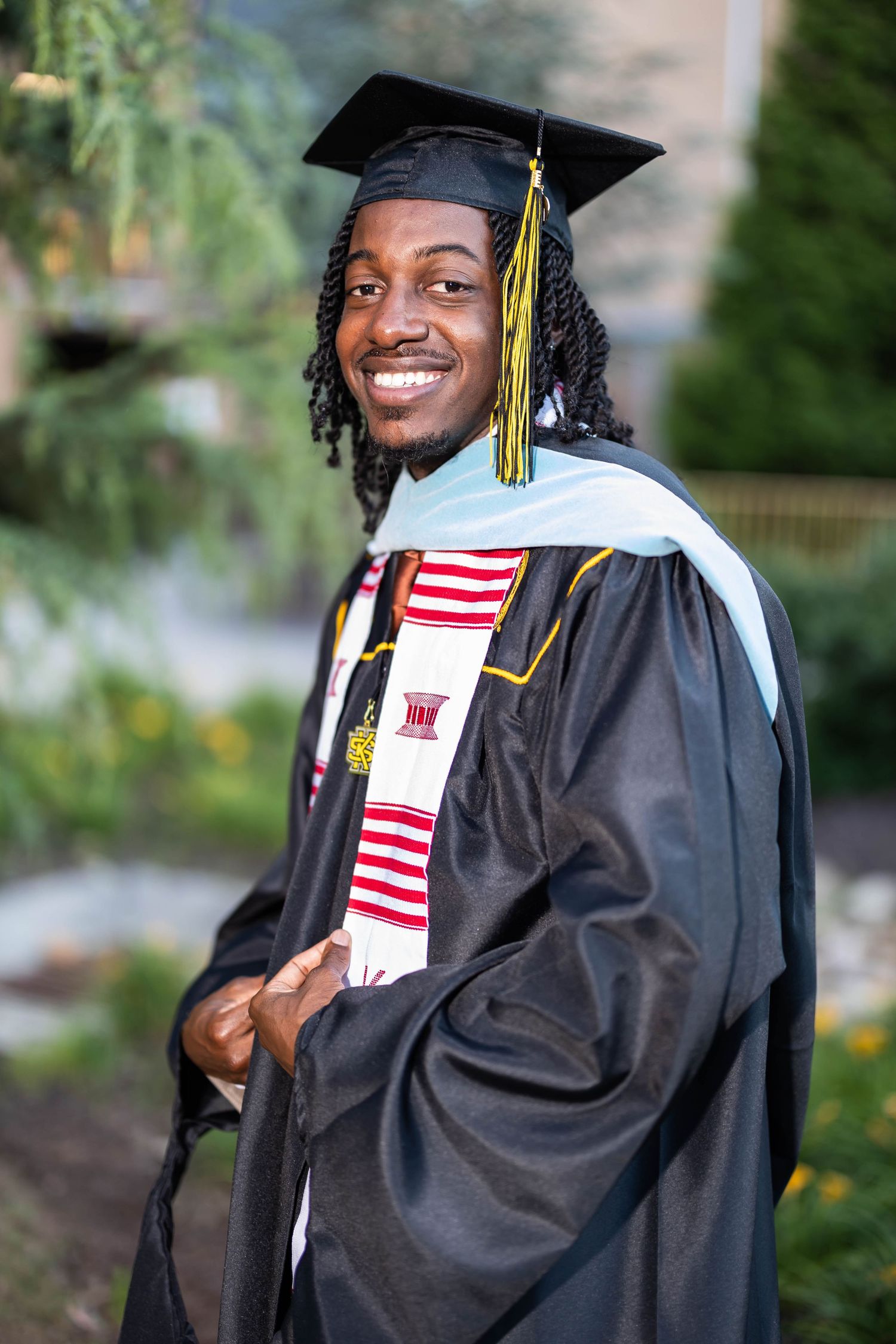 Smiling graduate in black cap and gown with kente stole poses outdoors on graduation day.