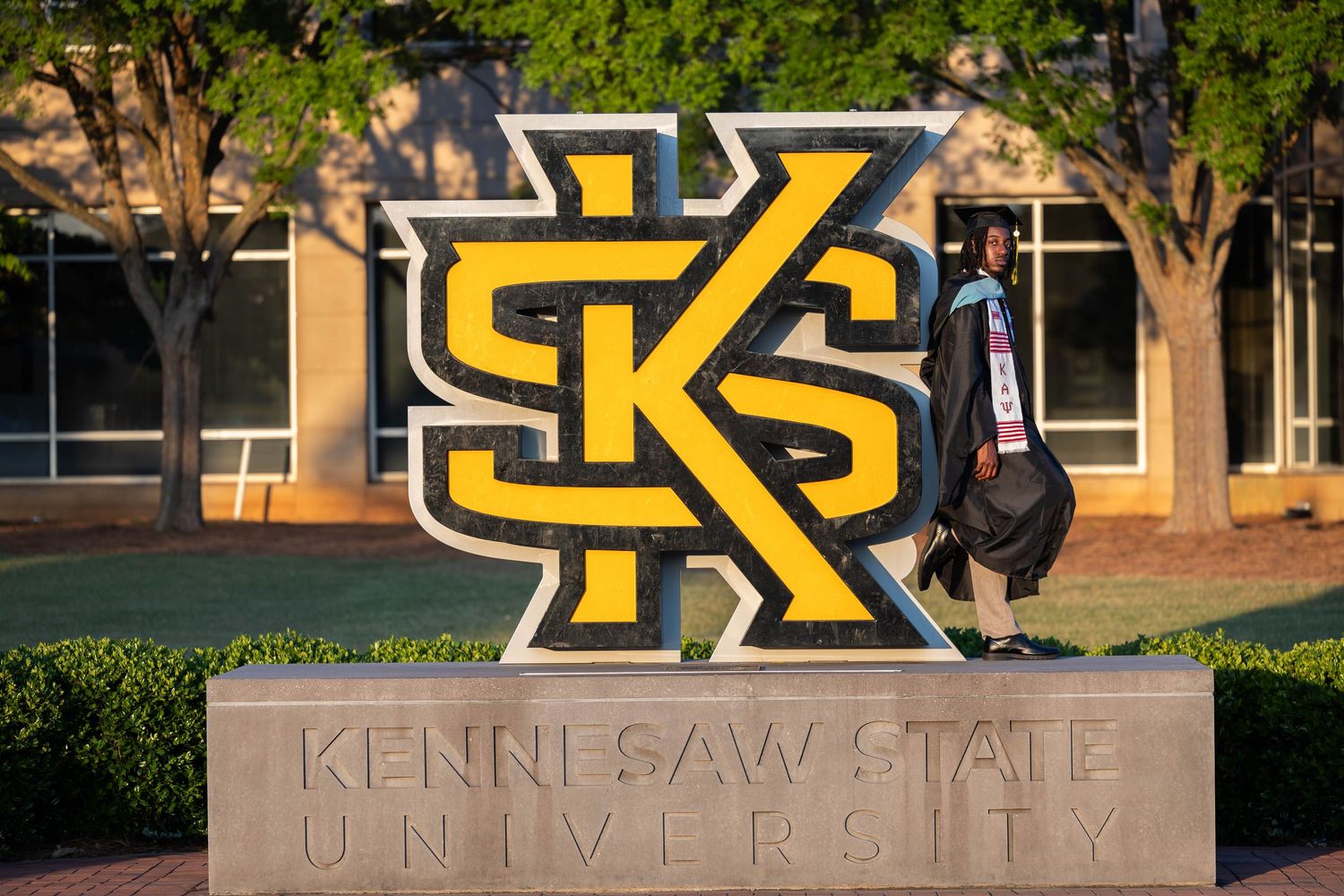 A graduate poses beside the iconic Kennesaw State University KSU logo sign on a sunny campus day.