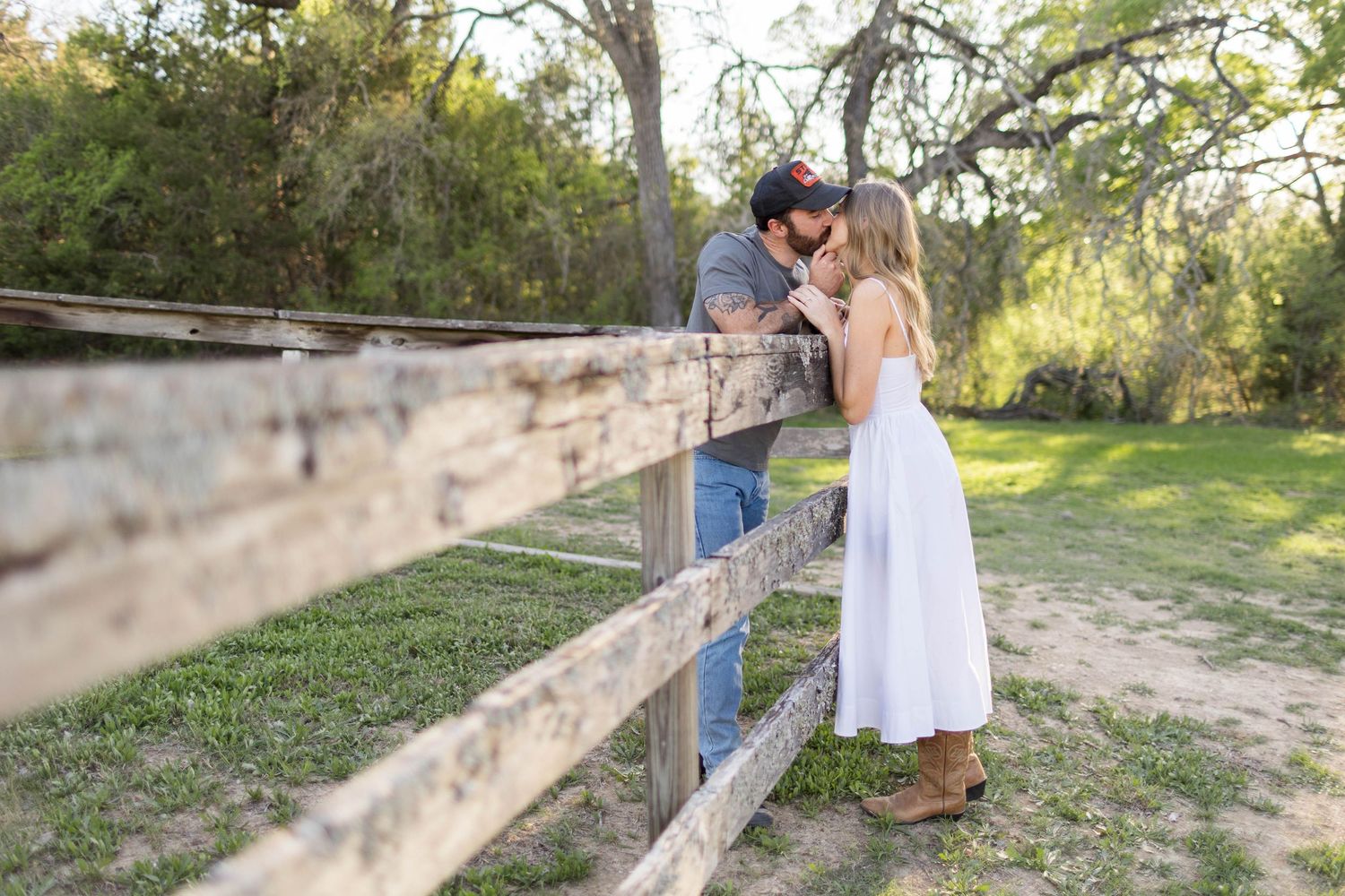 Couple sharing a romantic kiss over a rustic wooden fence in a lush green countryside setting captured by Austin engagement photographer.