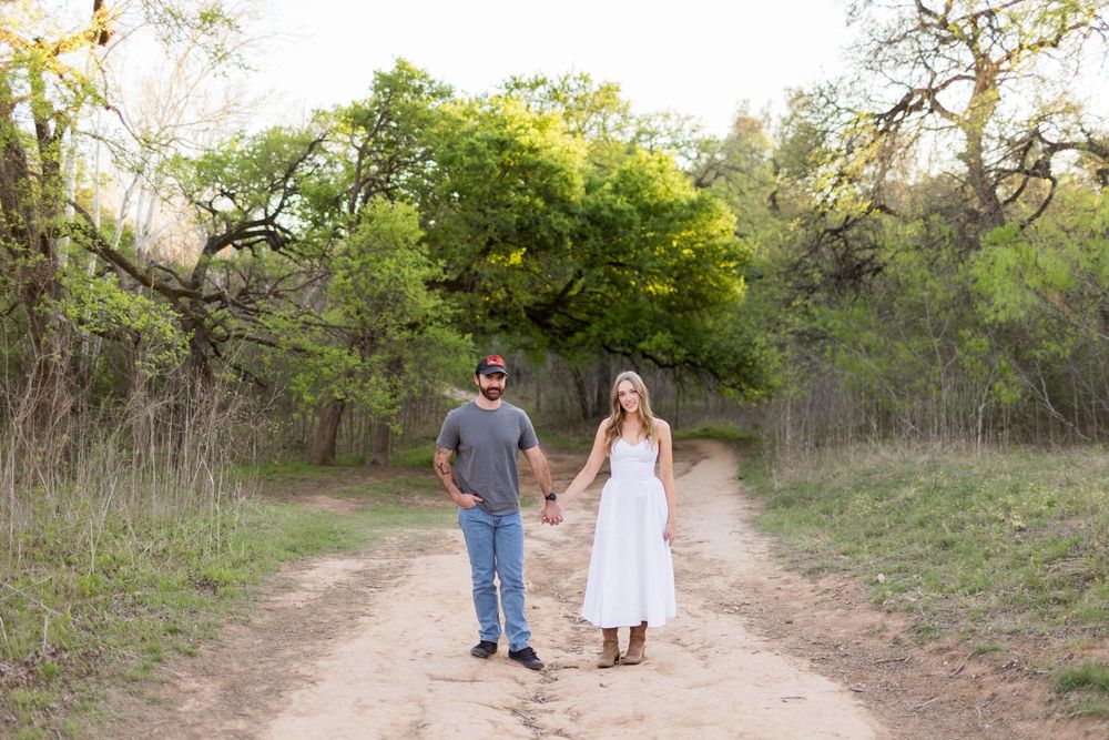 Couple holding hands on a dirt path surrounded by lush green trees during golden hour outdoor engagement photo.