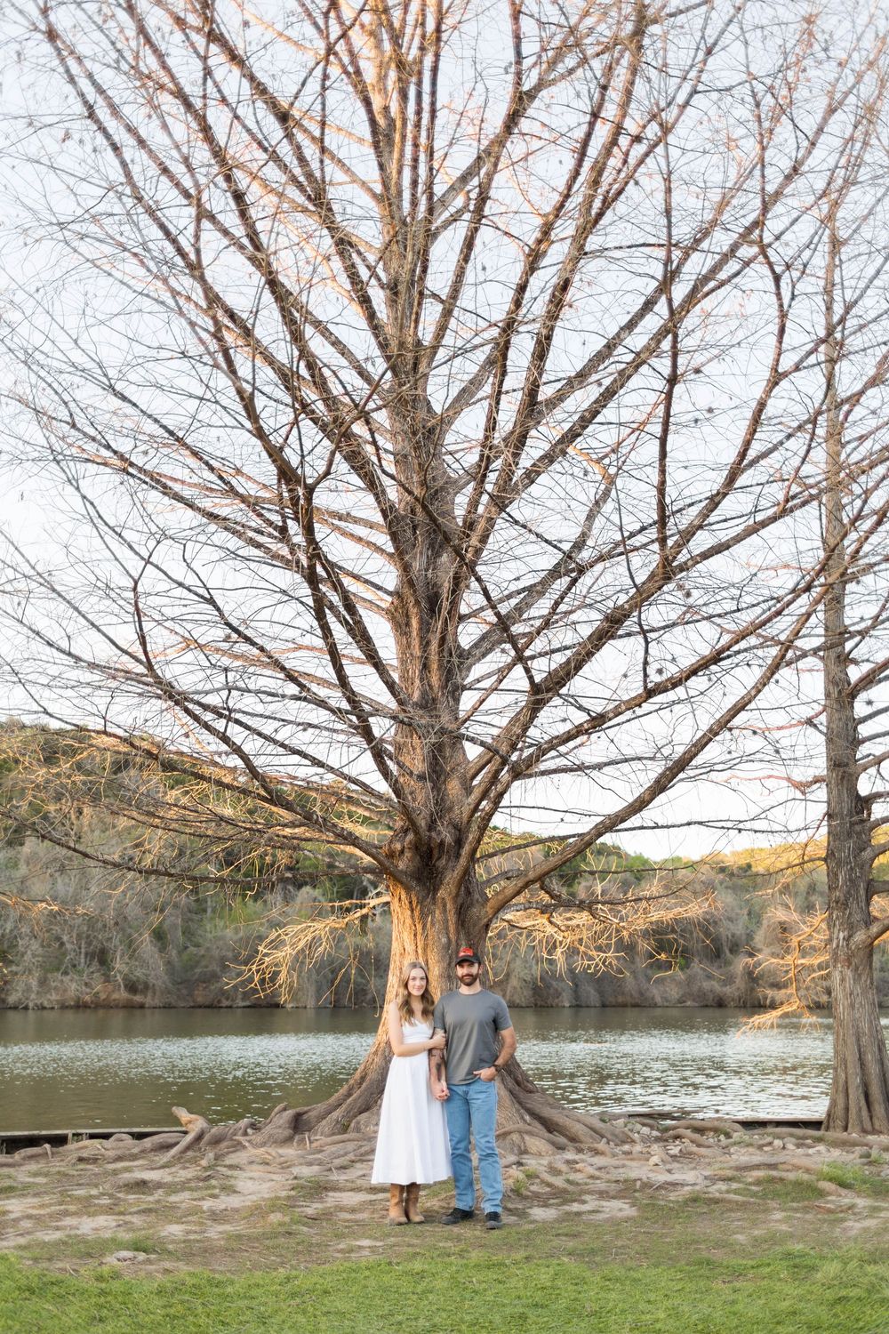 Couple standing together under a large bare tree beside a peaceful river in a scenic outdoor setting.