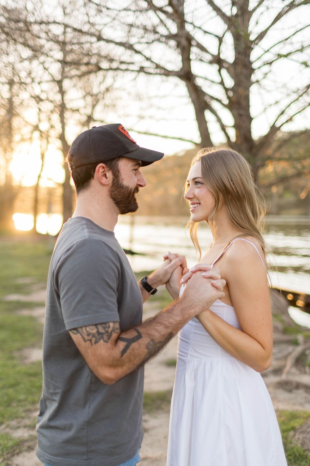 A couple holds hands and gazes at each other during a golden sunset by a lake, man in gray tee and cap, woman in white dress.